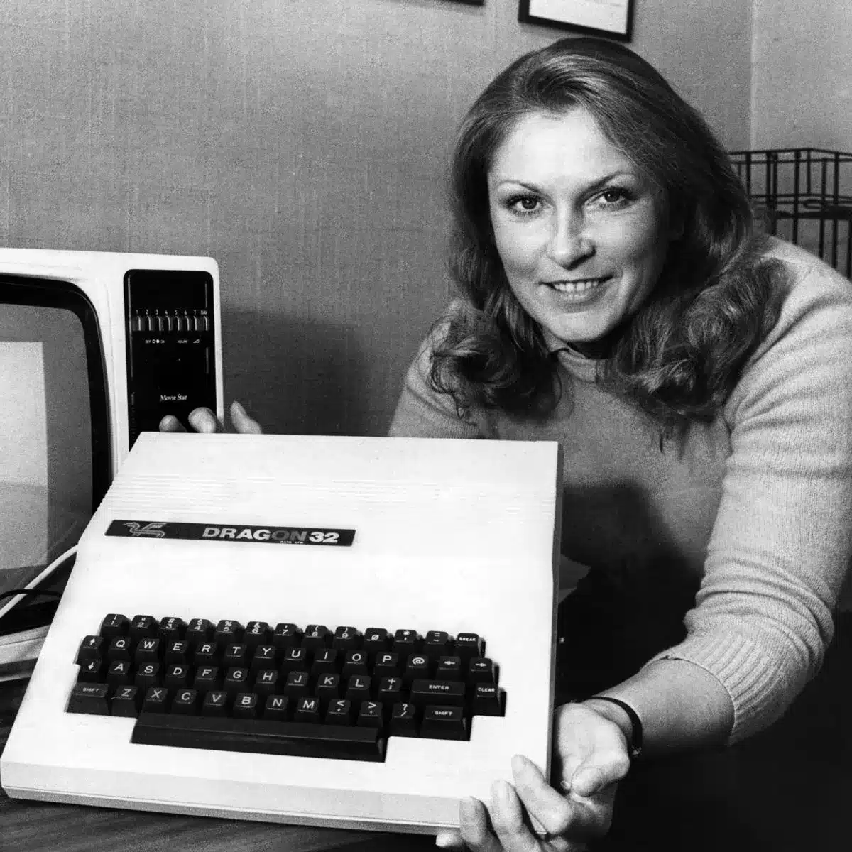 A period publicity photograph of a woman presenting a Dragon 32 alongside its monitor.
